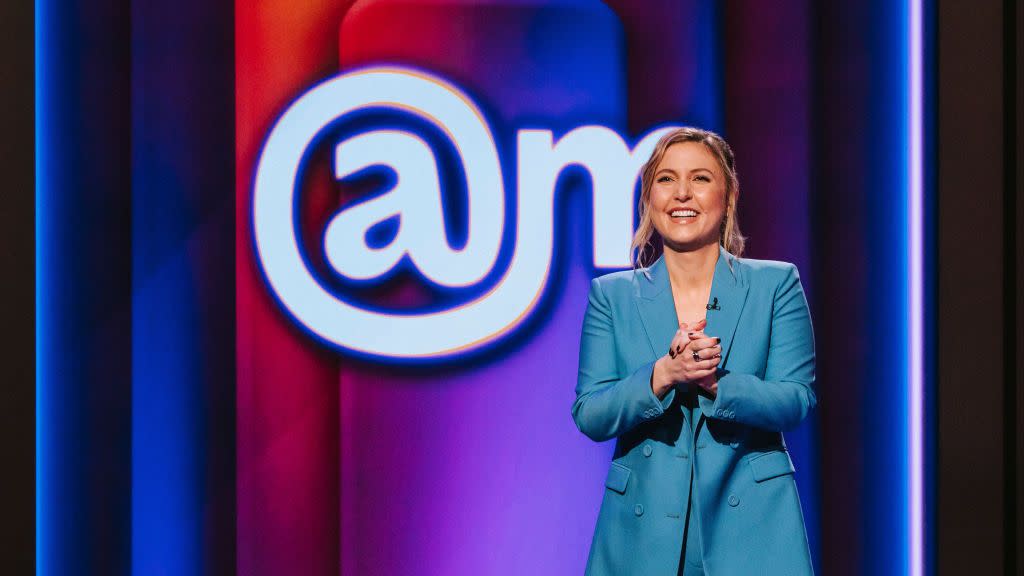 taylor tomlinson smiles while clasping her hands together in front of her chest, she stands in front of a multicolor background with the logo at sign am and wears a bright blue monochrome suit