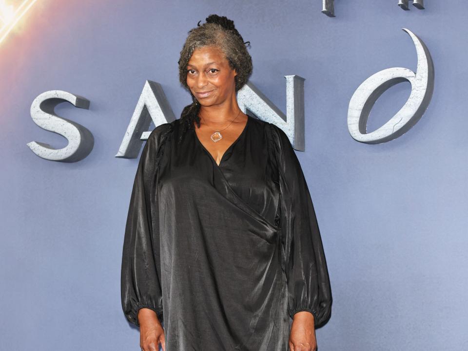 sandra james-young on the red carpet of the sandman, wearing a loose fitting, grey velvet dress. her hair is worn partially up, and she's smiling, looking slightly to the side