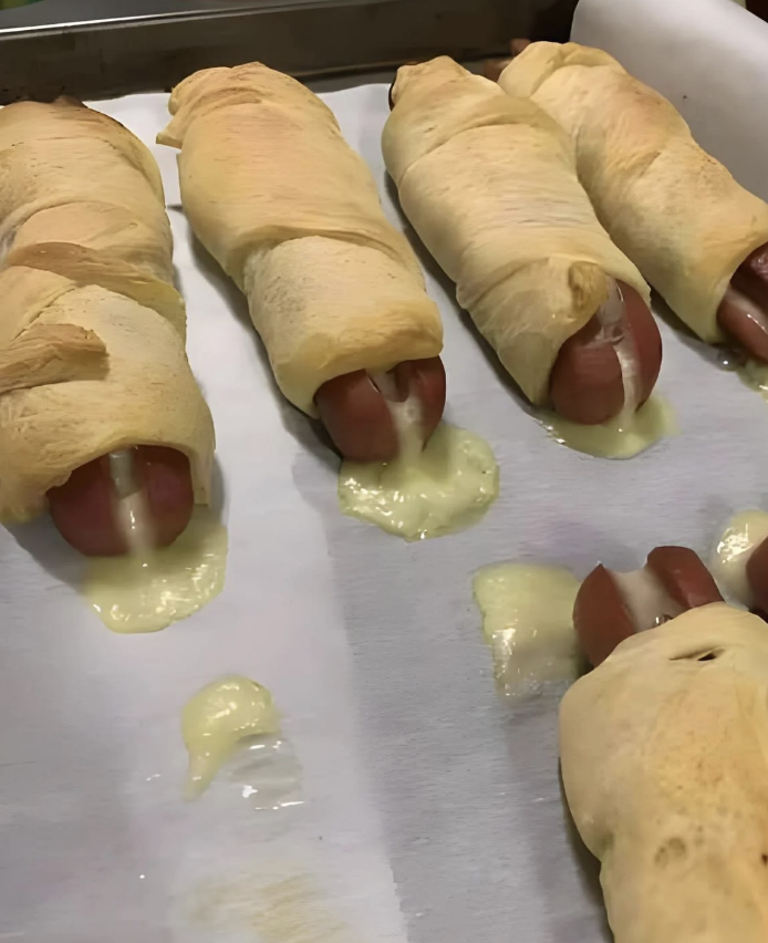 Hot dogs wrapped in puff pastry, with melted cheese oozing out of the sides on a baking tray