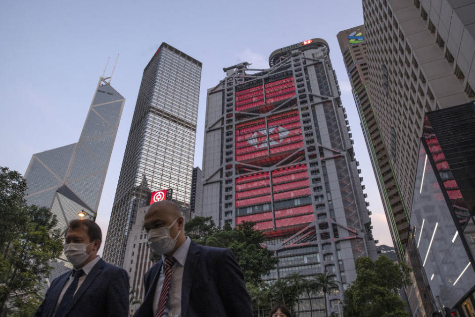 Pedestrians wearing protective masks walk as the HSBC Holdings Plc headquarters building, center, stands illuminated in the Central district of Hong Kong, China, on Monday, April 27, 2020. | Roy Liu/Bloomberg via Getty Images