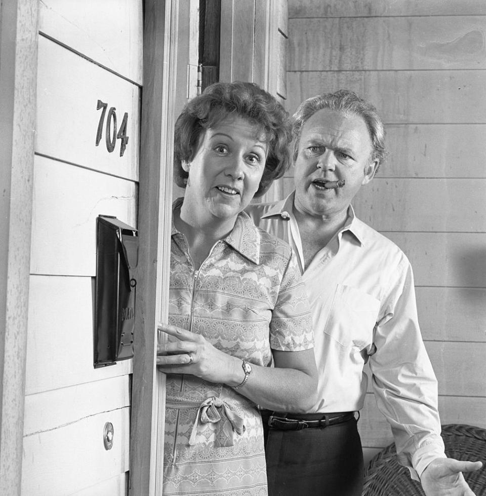 Jean Stapleton and Carroll O'Connor standing at the door labeled 704, appearing surprised. Jean Stapleton is wearing a patterned dress, while Carroll O'Connor is in a light shirt and dark pants