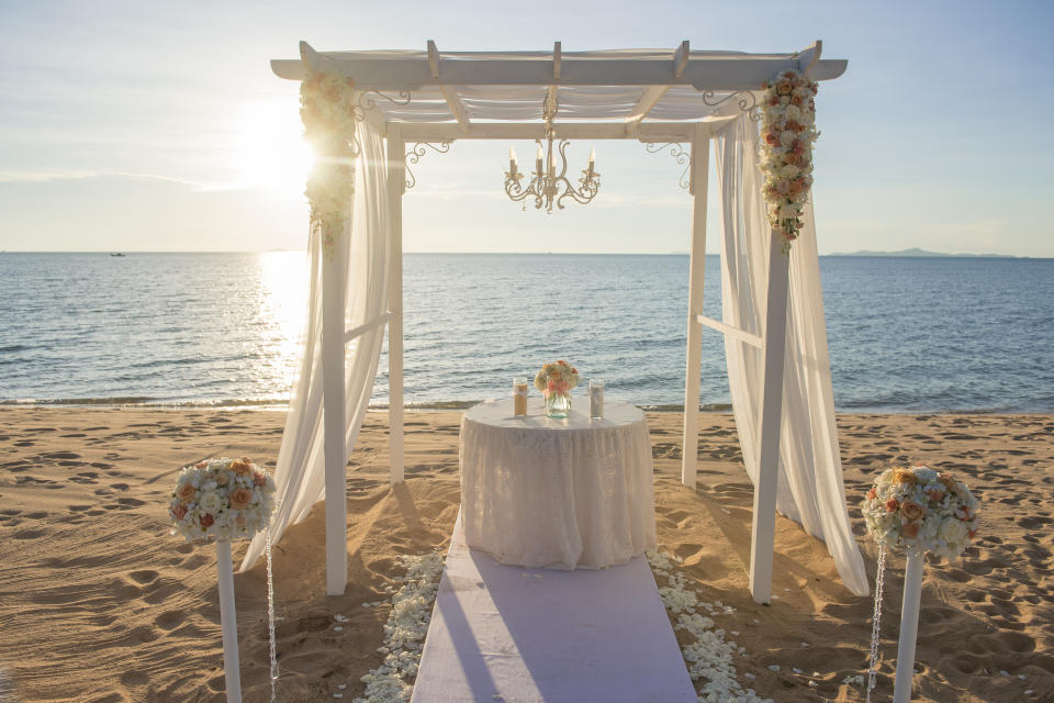 A wedding arch decorated with flowers and a small white table, set up on a beach with the ocean and sunset in the background