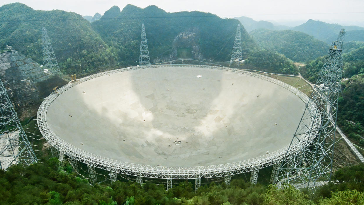  A large man-made dish sits like a crater surrounded by utility towers in a mountainous forest. 