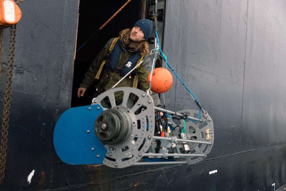 A man wearing a beanie stands inside a cruise ship with a giant piece of camera equipment about to be lowered into the water