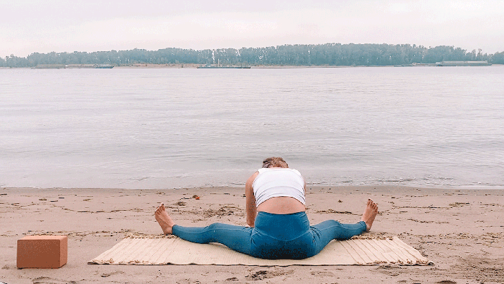 Woman sitting on a yoga mat at the beach practicing a Yin Yoga pose with her legs wide while leaning forward