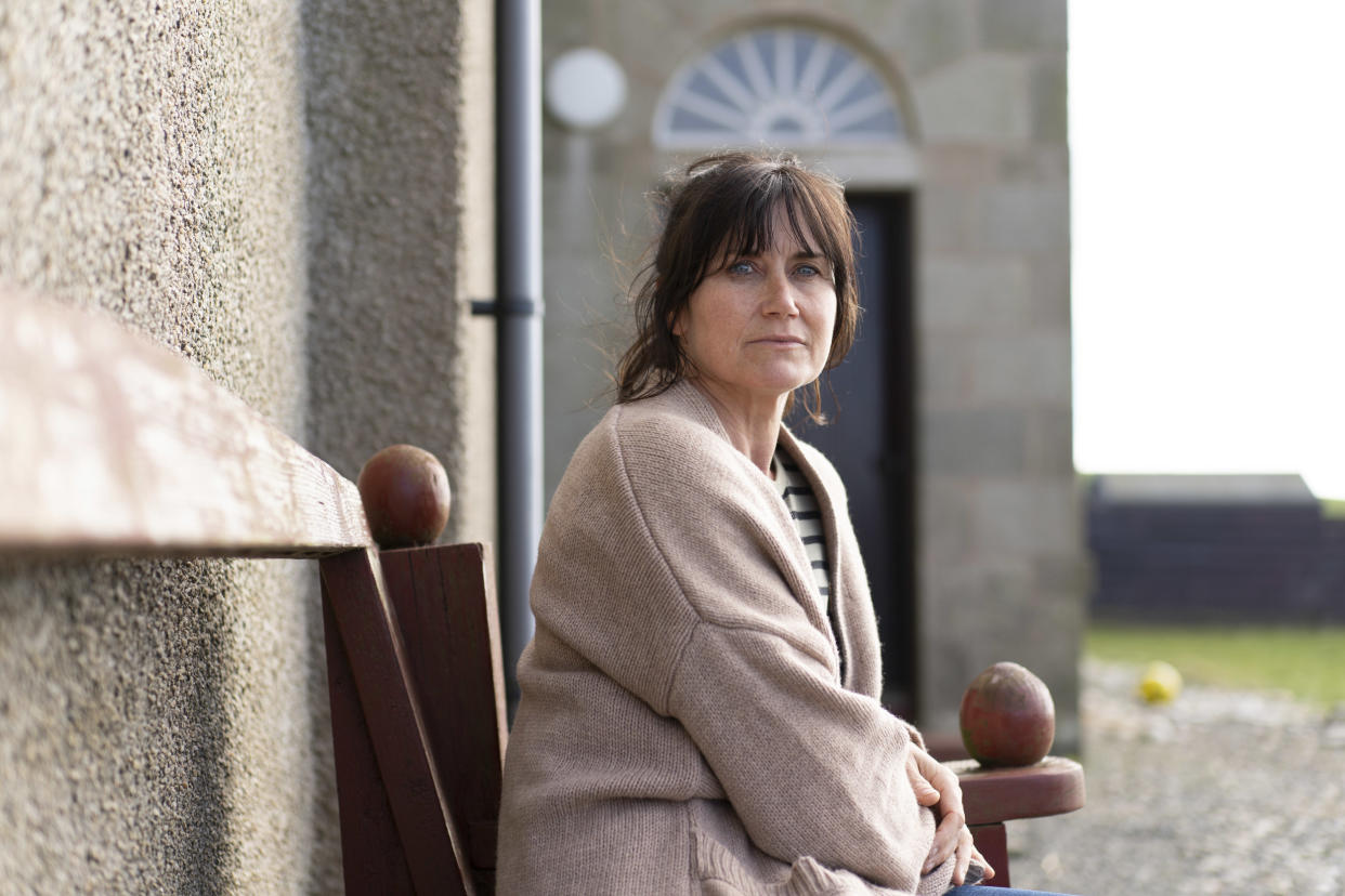  Shetland 's Stella Quinn (Dawn Steele) sits on a bench outside her family's house, with the front door behind her. She is wearing an oversized camel-coloured cardigan and jeans. 