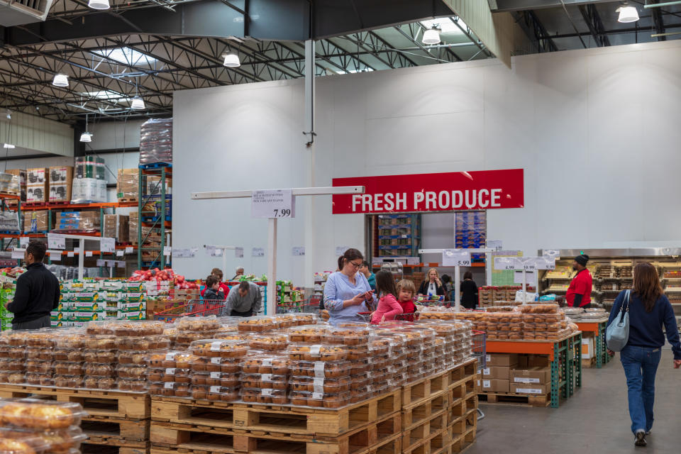 Shoppers at a warehouse store with "FRESH PRODUCE" sign, browsing items on pallets