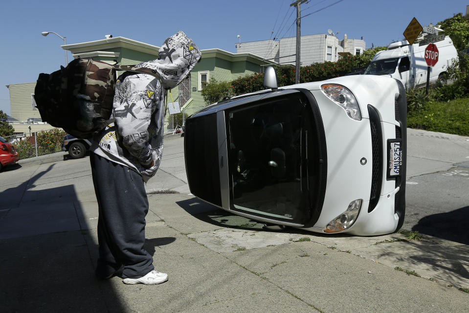 CORRECTS TO PROSPECT AND COSO AVENUES- A man looks at a tipped over Smart car on the corner of Prospect and Coso Avenues in San Francisco, Monday, April 7, 2014. Police in San Francisco are investigating why four Smart cars were flipped over during an apparent early morning vandalism spree. Officer Gordon Shyy, a police spokesman, says the first car was found flipped on its roof and a second was spotted on its side around 1 a.m., Monday in the Bernal Heights neighborhood. (AP Photo/Jeff Chiu)