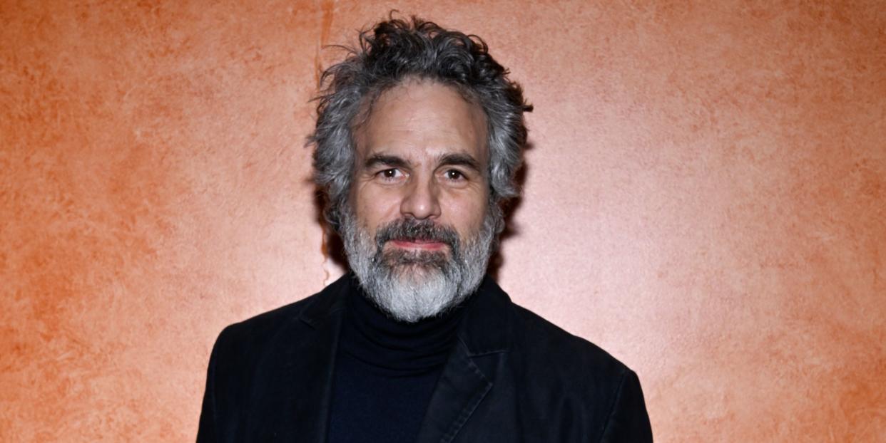 mark ruffalo, a man with gray and black hair and a gray and black beard, wearing black pants, a black coat, and a black turtleneck, poses in front of an orange wall