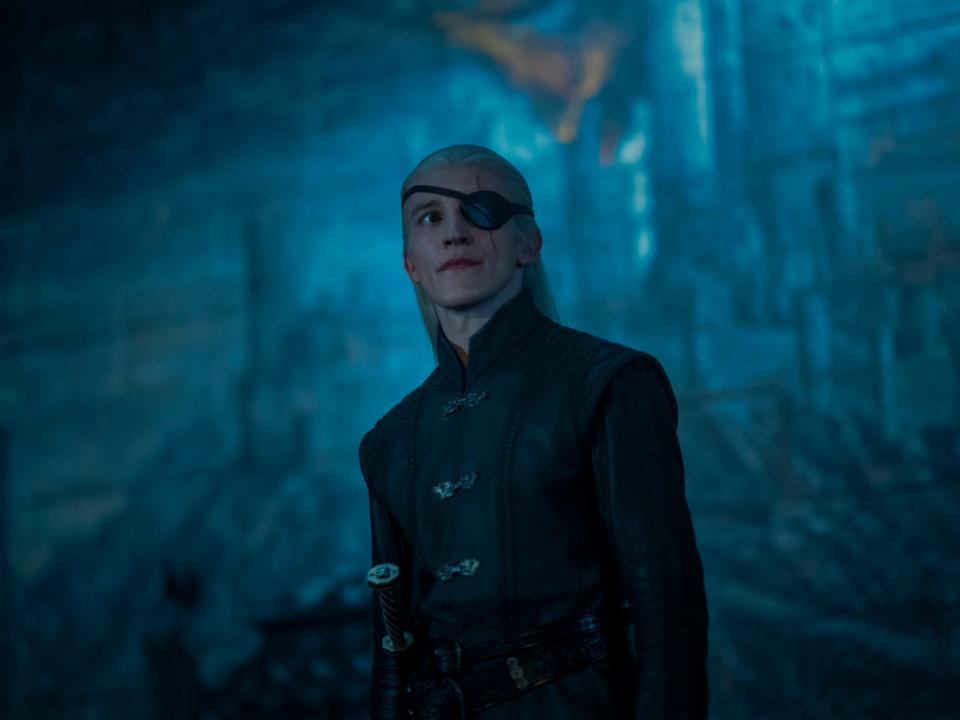 ewan mitchell as aemond targaryen, wearing black clothes, a sword at his hip, and an eyepatch over his left eye. he has long blonde hair pulled half back