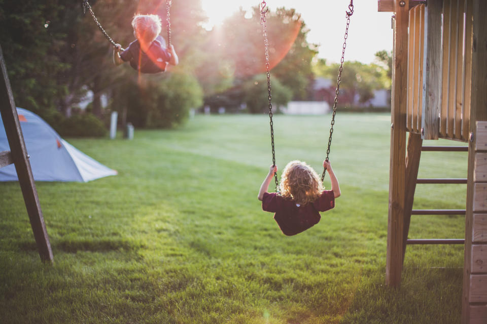 Back view of a little boy and little girl swinging high into the sky as the sun sets behind them while a tent is set up in the yard.