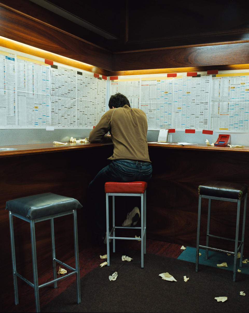 Person sitting at a high bar-like desk covered with papers on the wall, facing away from the camera. Crumpled papers are scattered around the floor