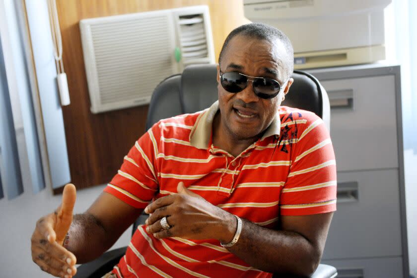 Saint Obi speaks and gestures while sitting in a chair and wearing sunglasses and a striped red shirt.
