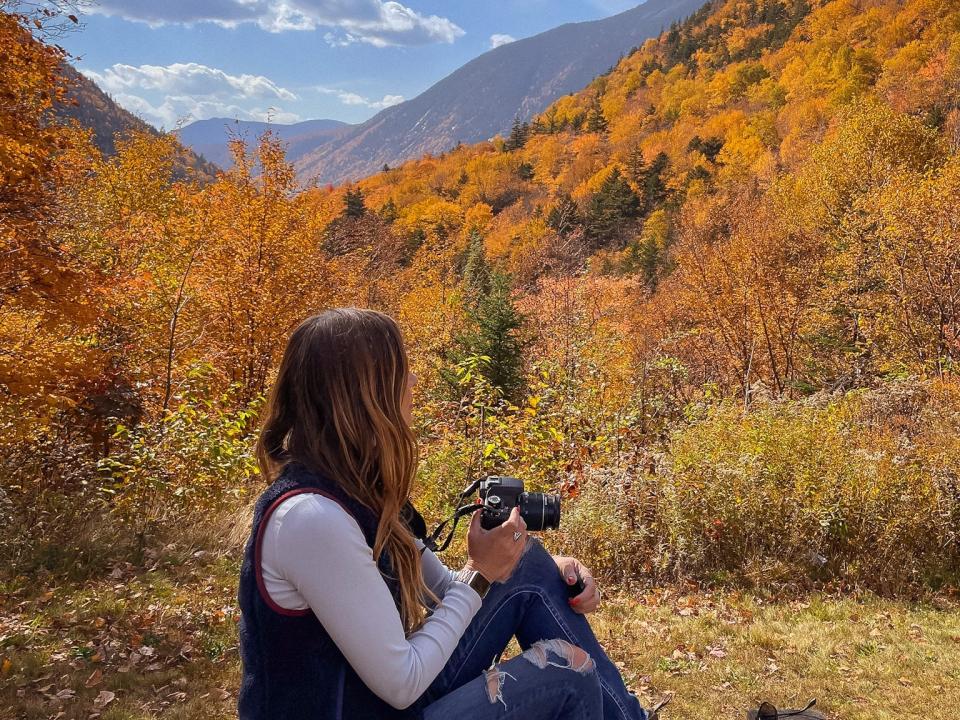 Emily sits on a wall facing to the right with a camera in her hand. Behind her are mountains and trees with beautiful fall foliage.
