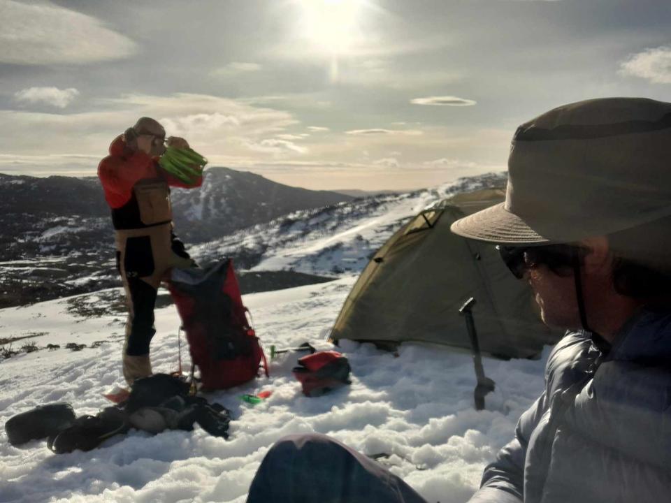 Two men on the top of an Australian mountain that's covered in snow. We can see a tent behind them. The sun is bright and high in the sky.