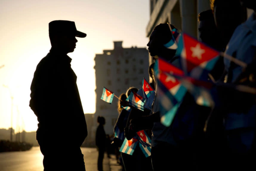 <p>Die Silhouette eines Soldaten zeichnet sich vor dem Morgenhimmel ab. Menschen tragen kubanische Flaggen und warten auf die Autokolonne, die den Sarg des kubanischen Präsidenten Fidel Castro in Havanna, Kuba, transportiert. (Bild: AP Foto/Natacha Pisarenko) </p>