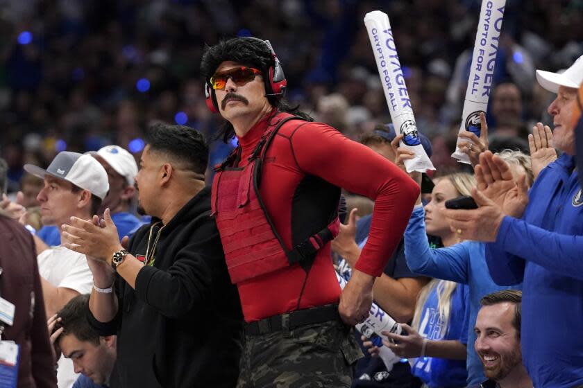 A man with a dark mustache, a dark mullet and sports sunglasses in a red longsleeve shirt standing amid sports fans in blue