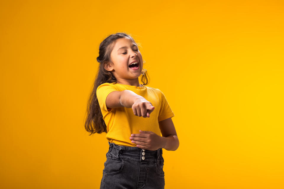 Portrait of kid girl mocking and teasing at someone showing finger at camera and holding stomach over yellow background. Bulling concept