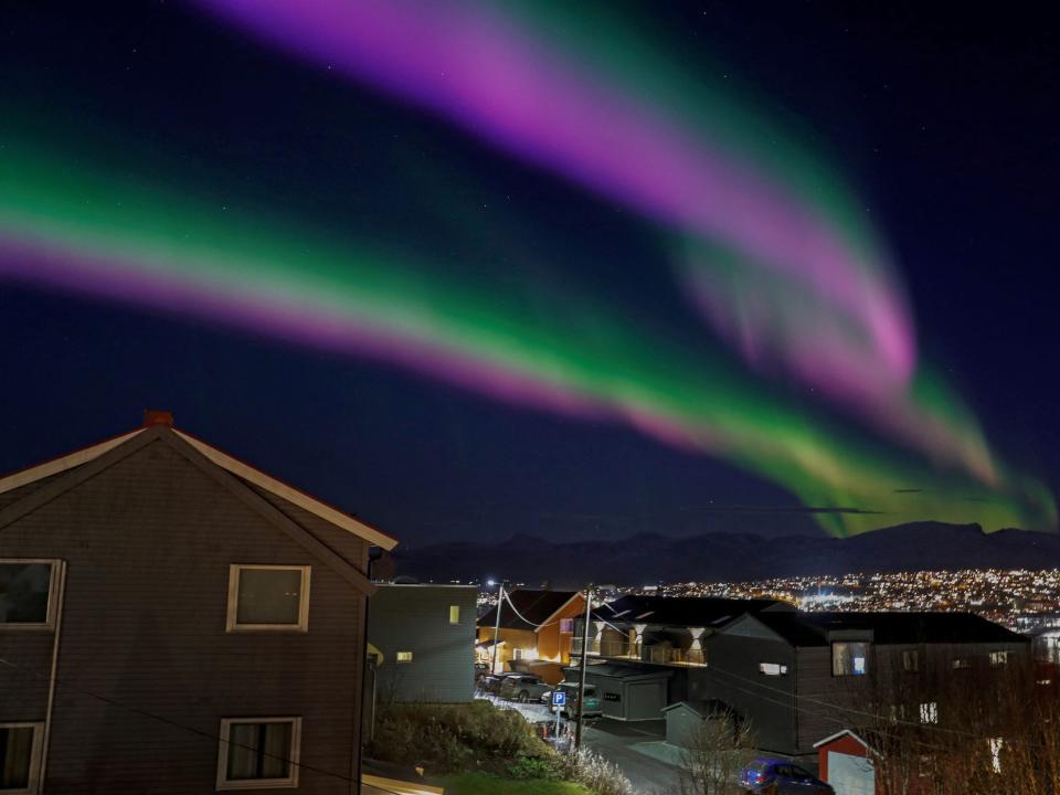 ribbons of green pink purple northern lights stretch across the night sky above homes and lights of a sprawling town