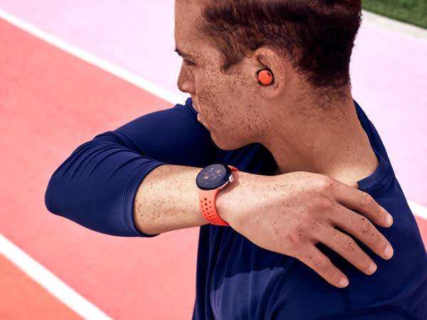 Promotional image of an athlete wearing a dark blue t-shirt wearing a matching Pixel Watch 2 coral band and buds.