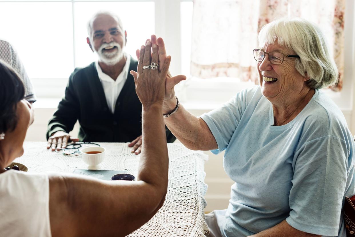 Two senior women giving each other a high five around a table with a doily tablecloth with two other seniors, both males, with a bright window blurred in the background in a living room