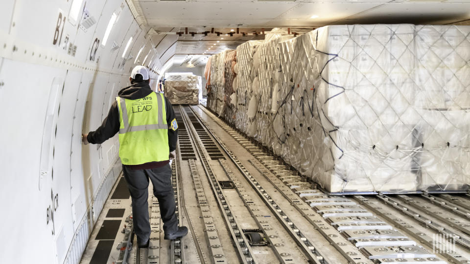 Man in yellow vest monitors loading of pallets on roller system inside body of aircraft.