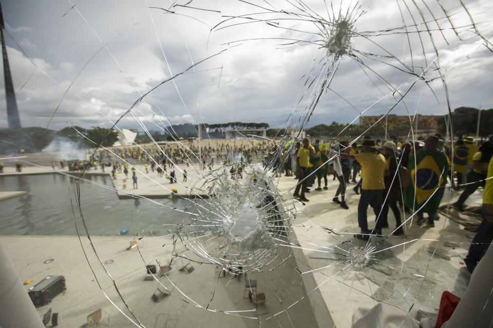 Seen through cracked glass, hundreds of people, many of whom wear yellow and green shirts, stand on a concrete park next to a small pond.