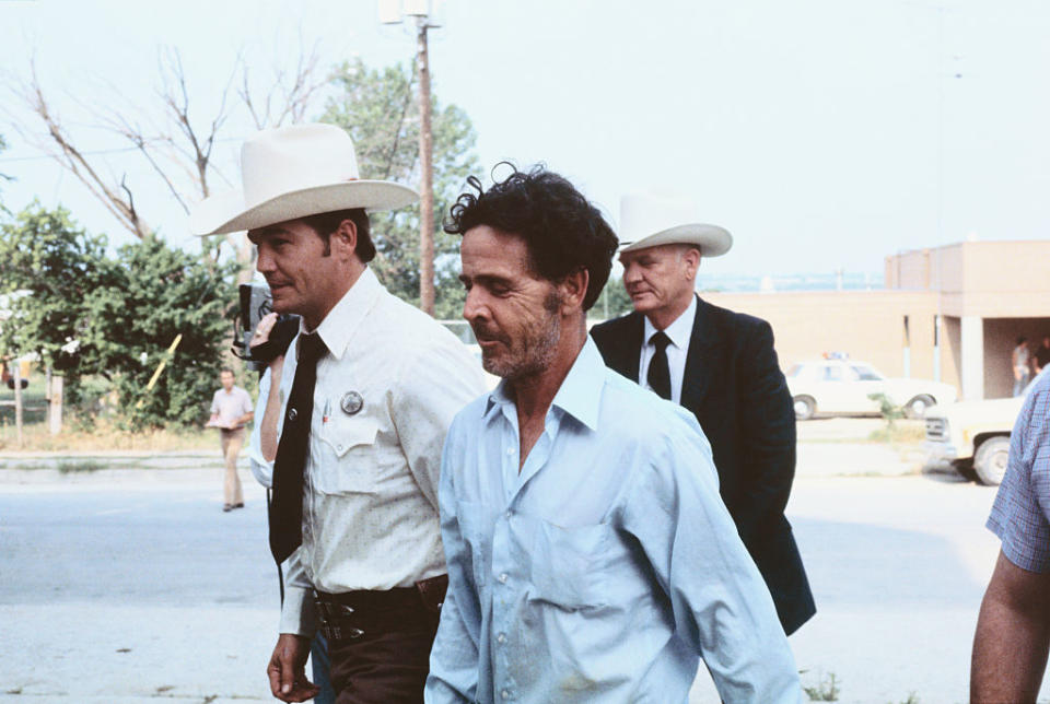 Three men, including Woody Harrelson (center), dressed in casual and formal attire, are walking outdoors beside a law enforcement officer and another man in a cowboy hat