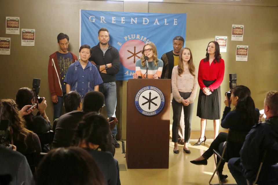 A group of seven community college students stand behind a podium, in front of a crowd. One woman talks into the podium's microphone.