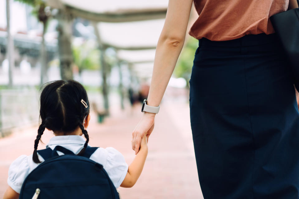 Rear view of young Asian mother taking her little daughter to school, they are holding hands and walking in the city street in the morning