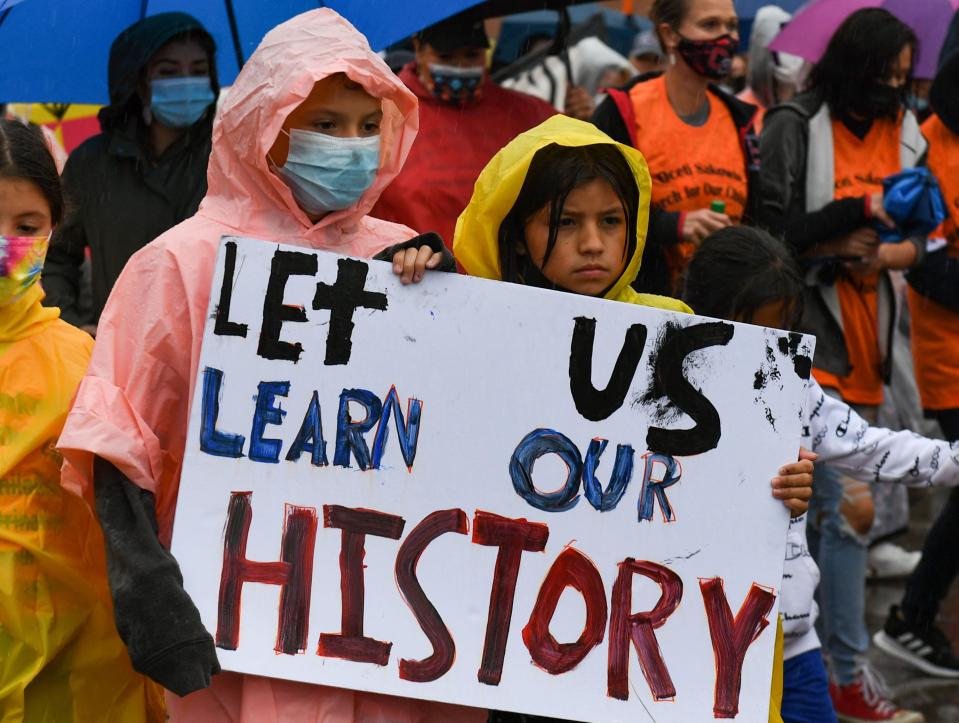 Jaylee Fallis and L. In The Woods march with a sign that reads "Let us learn our history" during a demonstration through the rainy streets of Pierre on Monday, September 13. More than 200 marchers walked, drummed and rode horses to demand Indigenous history education for all after the Dept. of Education removed more than a dozen references to the Oceti Sakowin before releasing proposed social studies standards a month ago.