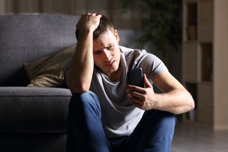 A young man sitting on the floor against a sofa, looking at his phone with a disappointed and confused look on his face