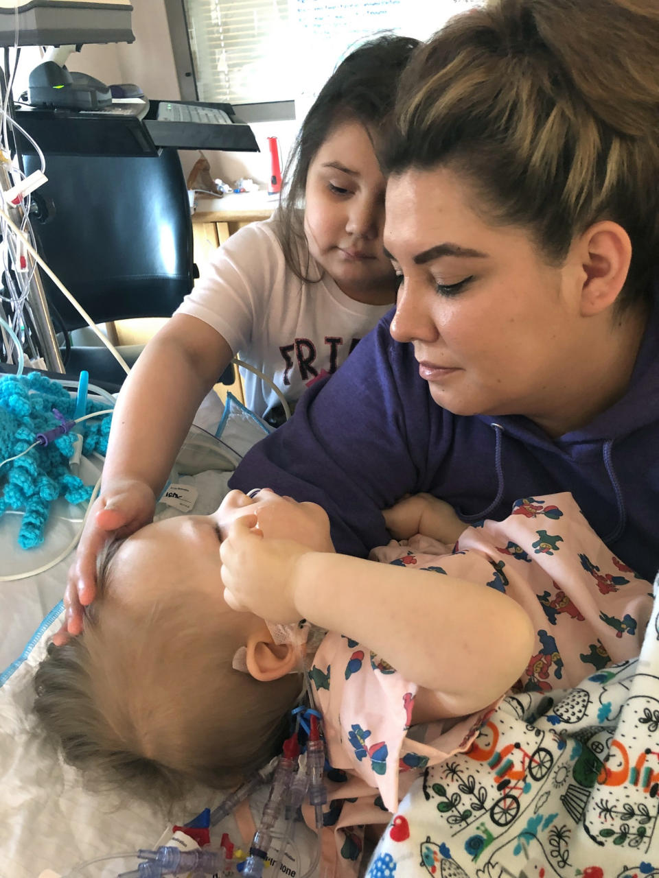 In this photo provided by Joan Azure shows Greyson Parisien, left, is comforted by his mother Reeanne Parisien and sister, Parker, at the Mayo Clinic in Rochester, Minn., in April 2019. Greyson's journey to correct a heart defect led the Turtle Mountain Band of Chippewa Indians to designate a spot on tribal IDs for organ donation. (Joan Azure via AP)