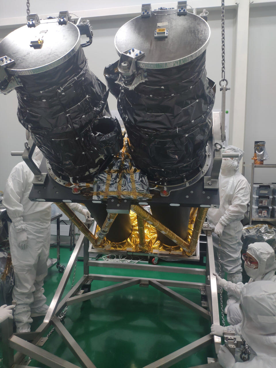 Three people dressed in full-body white jumpsuits work around a spacecraft shaped like two large black cylinders supported by a metal frame.