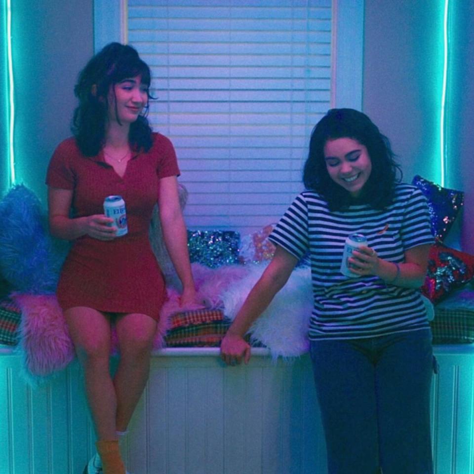 Two women, one in a red dress and the other in a striped shirt, are smiling and holding cans, seated in a neon-lit room