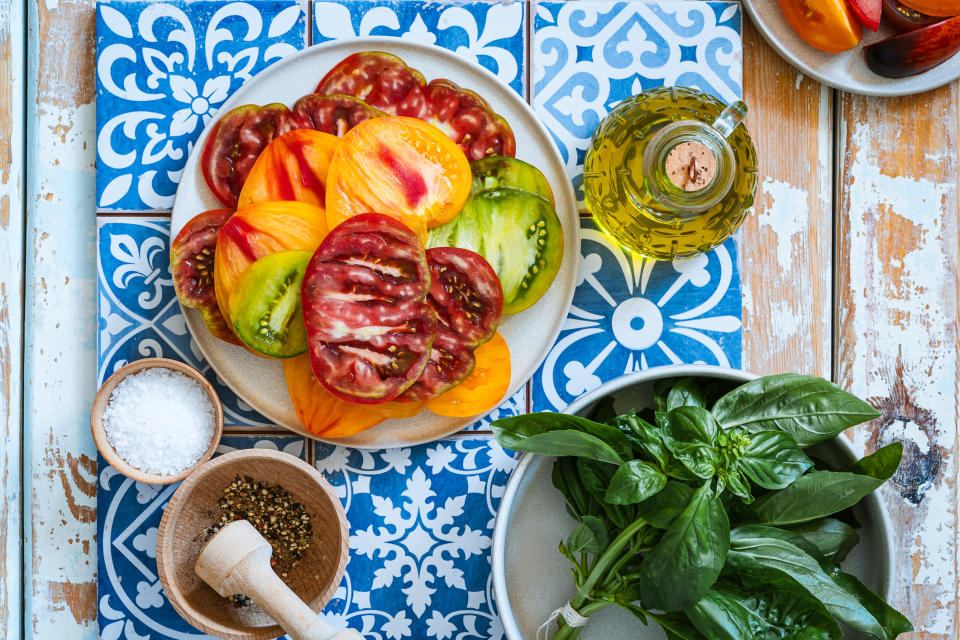 A plate of sliced heirloom tomatoes is surrounded by a small bowl of pepper, a bowl of salt, fresh basil leaves, and a bottle of olive oil on a decorative table