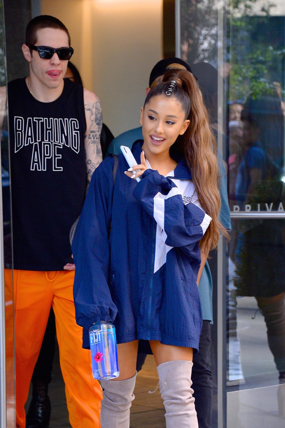 Ariana Grande, wearing an oversize men's jacket, walks out of a building waving, Pete Davidson is walking behind her