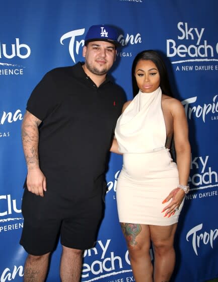 A man in a black polo shirt and shorts puts his arm around a woman in a white dress