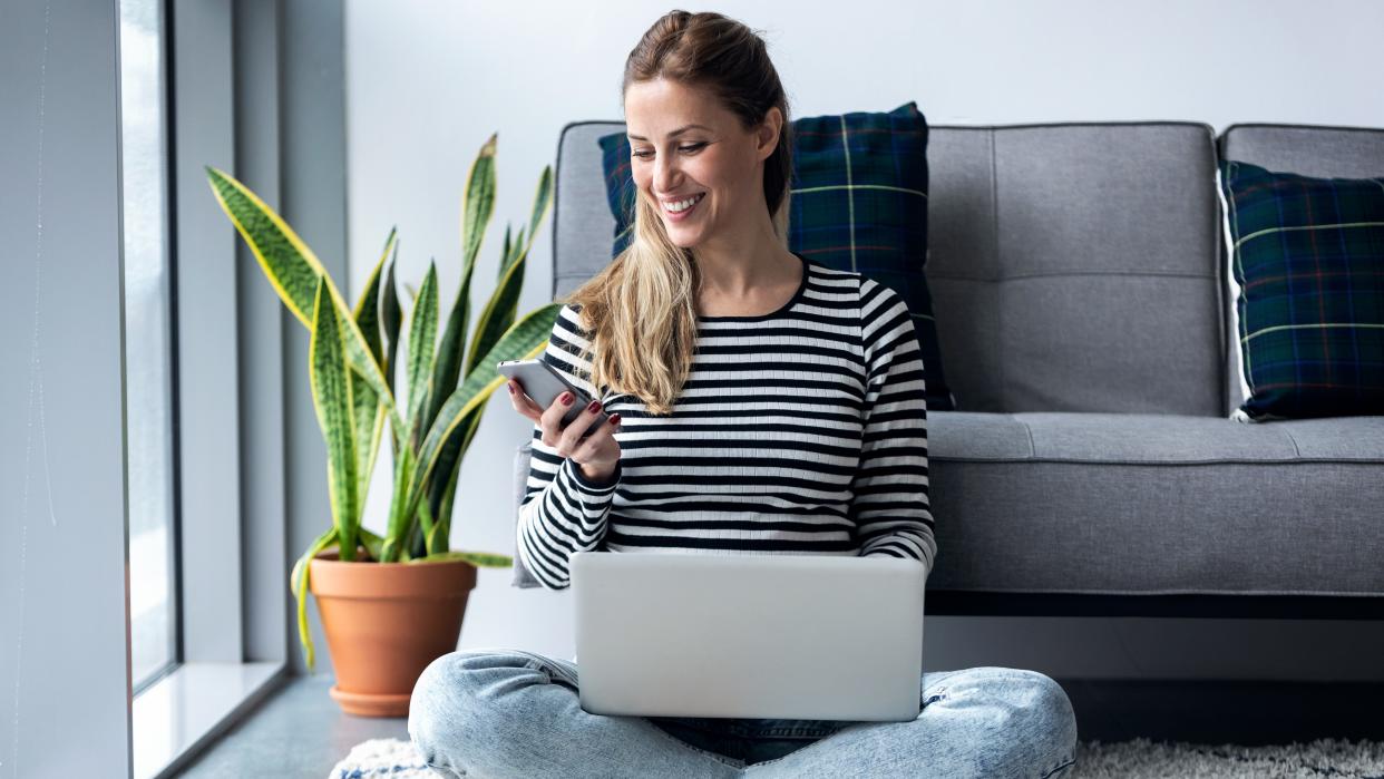   woman using her mobile phone while working with laptop sitting on the floor at home. 