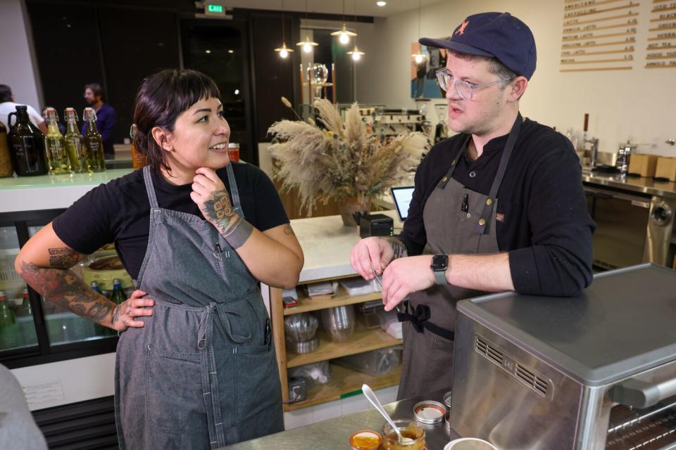 A woman and a man in chef's aprons stand behind a counter in a restaurant, talking.