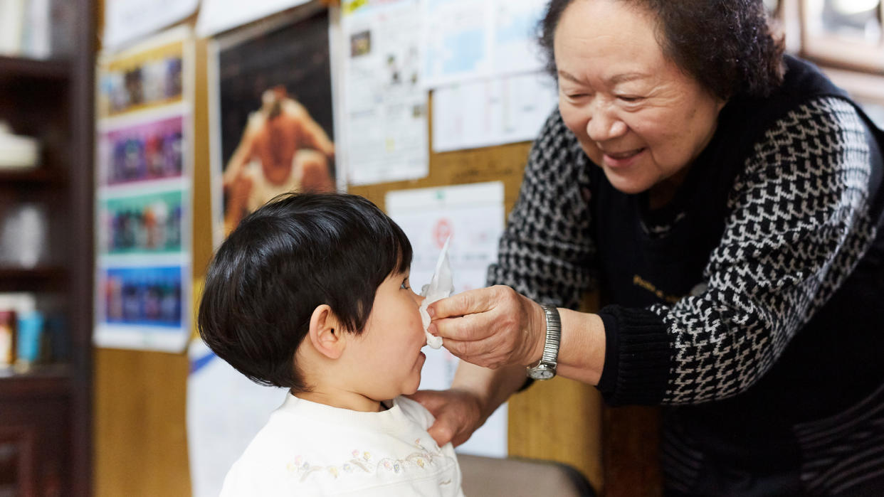  A grandmother smiles as she pinches a tissue over her young grandchild's nose; both are standing in what looks like a store with posters on the walls. 