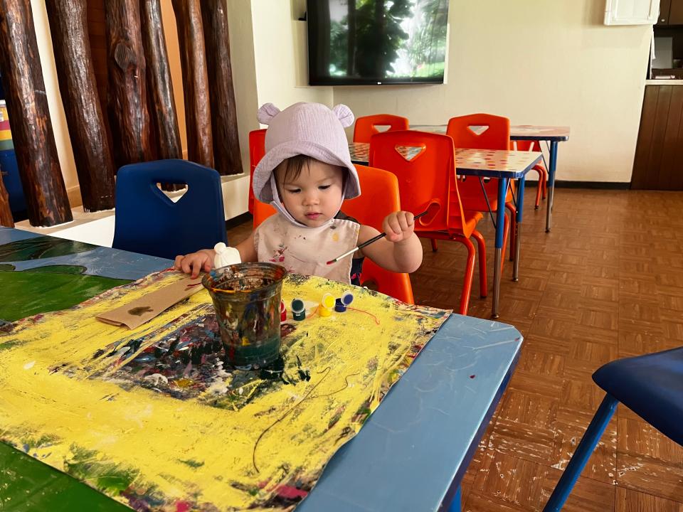 A child wearing a pink hat holds a paintbrush and paints on a yellow piece of paper