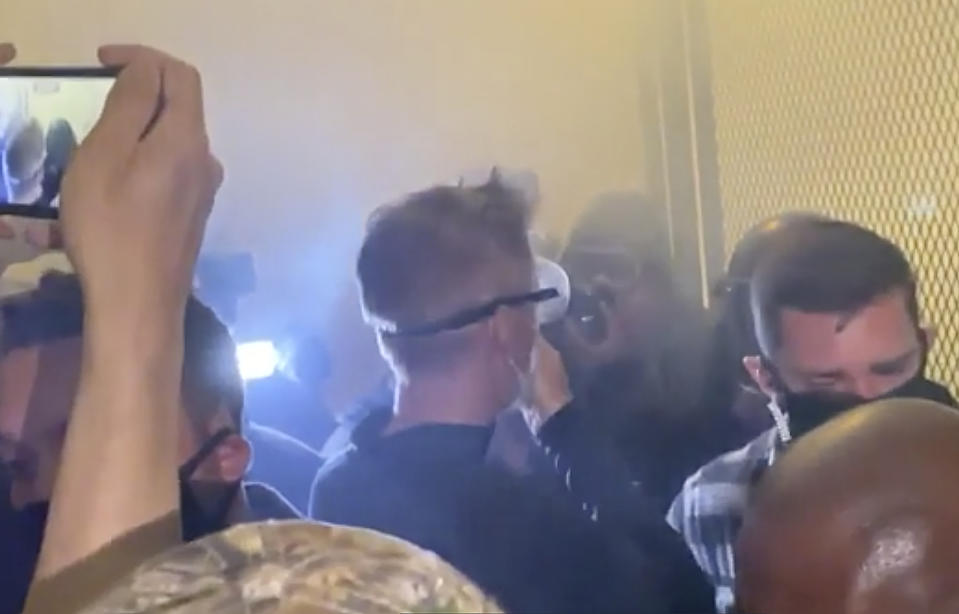 In this image made from video released by Jonathan Maus, Portland Mayor Ted Wheeler, center in black with goggles looking away, stands at a fence guarding a federal courthouse as tear gas drifts by early July 23, 2020, in Portland Oregon, during another night of protest against the presence of federal agents sent by President Donald Trump to quell unrest in the city. (Jonathan Maus/BikePortland via AP)