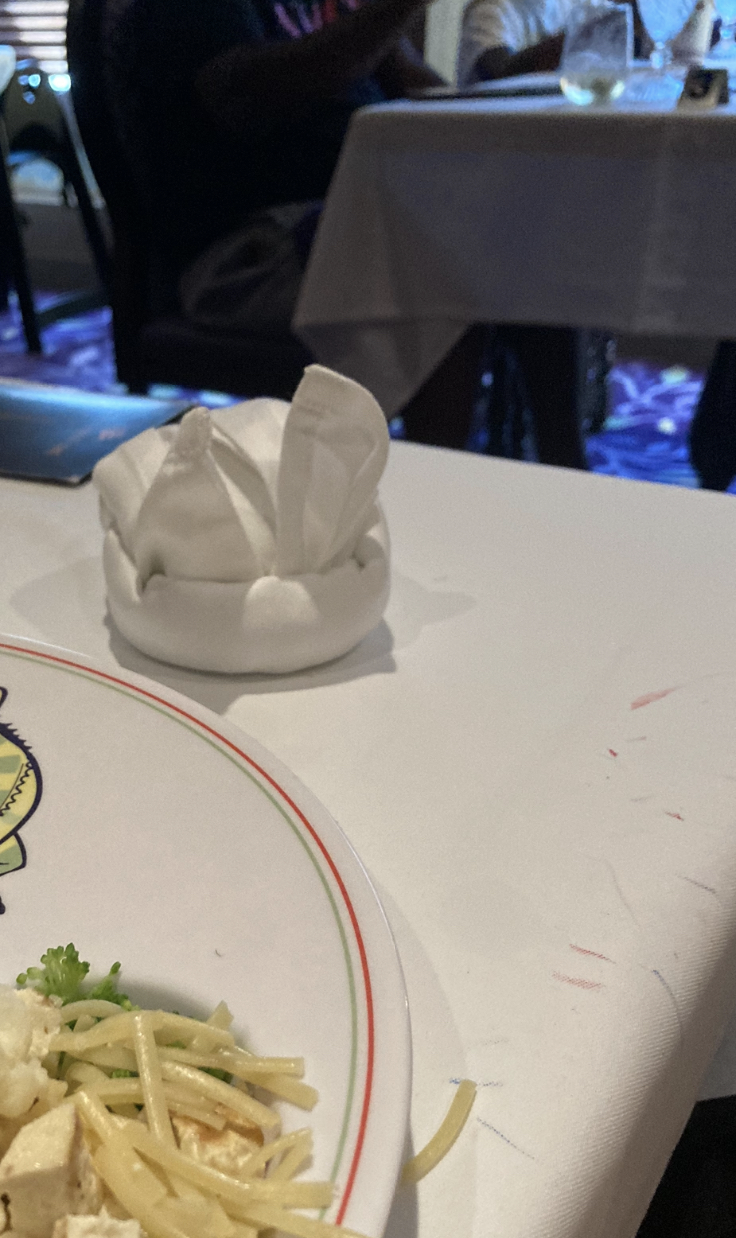 A table with a partially eaten plate of pasta and broccoli in the foreground. A cloth napkin folded creatively sits in the background