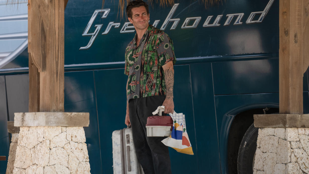  Jake Gyllenhaal stands smiling in front of a Greyhound bus in Road House. 