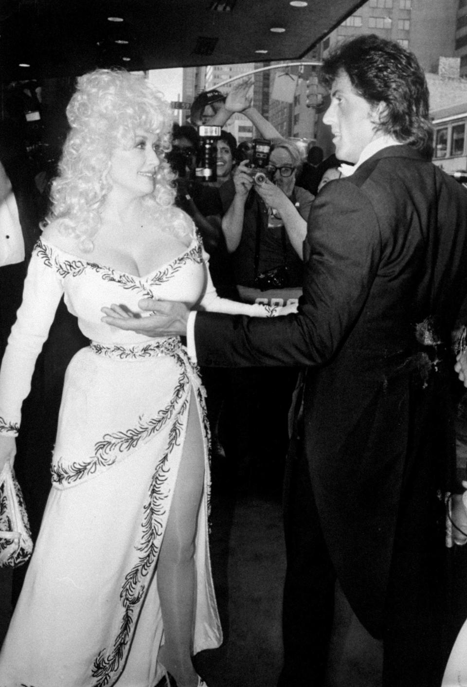 Dolly parton in a low cut white cross-body dress with Sylvester Stallone in a tuxedo.