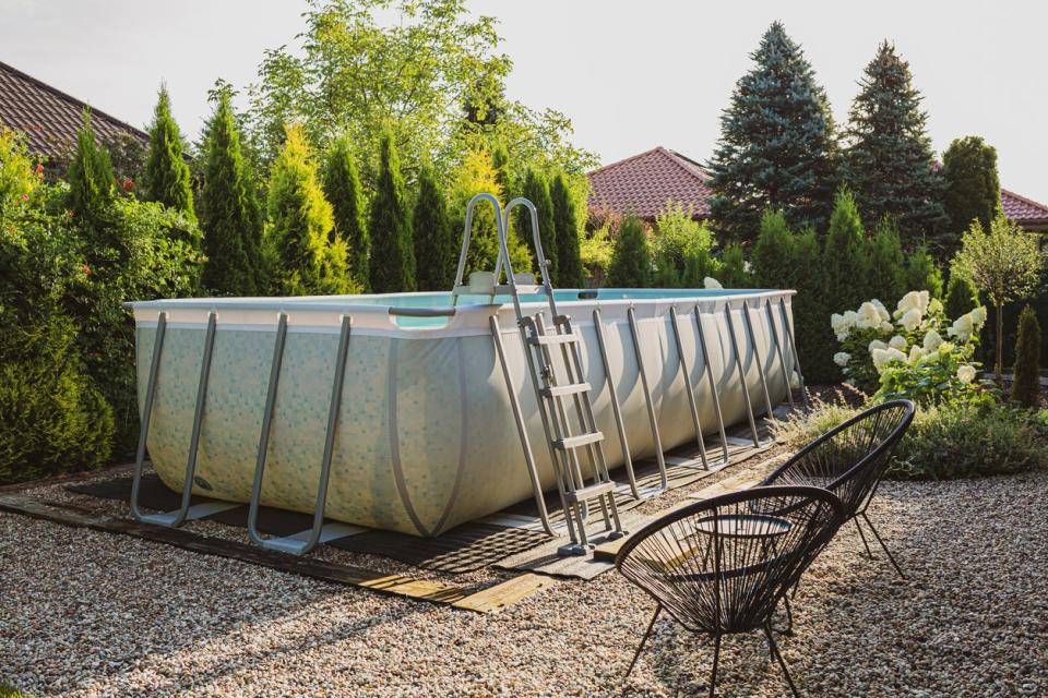 Rectangular above-ground swimming pool on gravel surrounded by trees, shrubs, and flowers