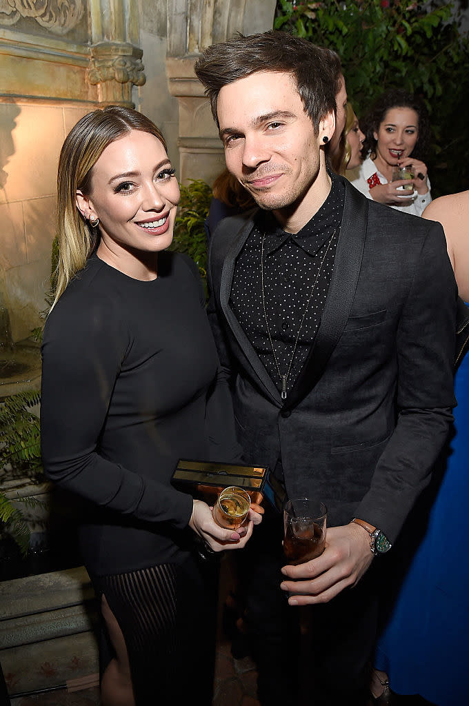 the couple smiling, Hilary in a long-sleeved dress and Matthew in a blazer with a dotted shirt, holding drinks at an event