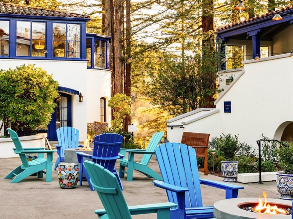 Blue chairs around fire pits on a porch beneath a white building with blue accents and greenery in the background