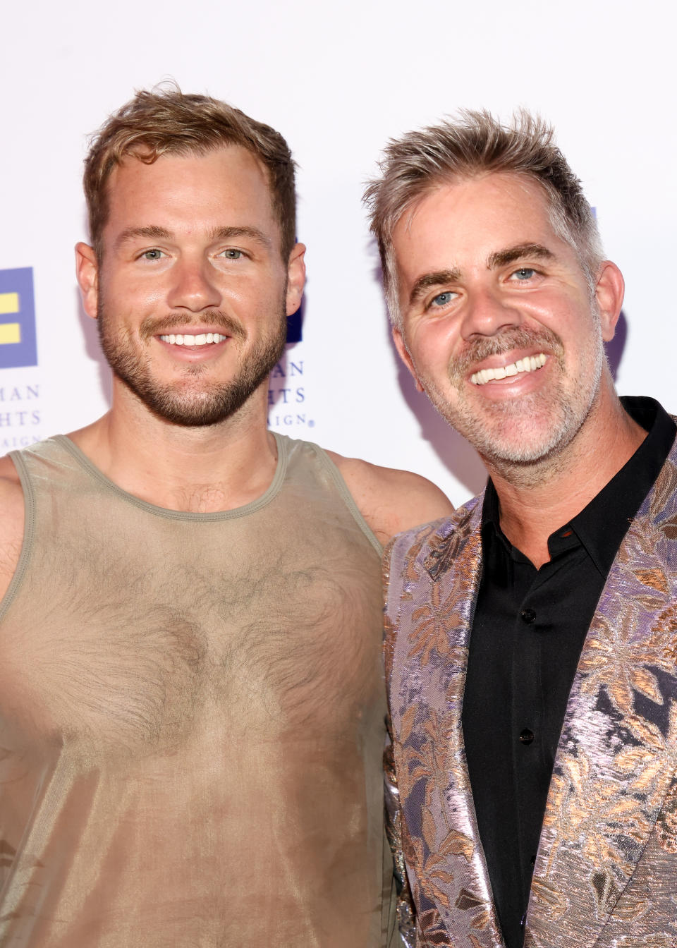 Colton Underwood and Jordan C. Brown pose together at an event, with Colton wearing a sheer top and Jordan wearing a shiny patterned blazer